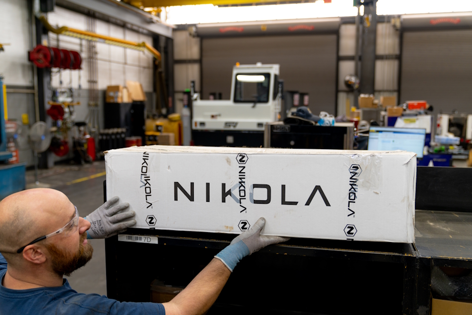 A worker wearing safety glasses in a workshop holding a box with Nikola on it.