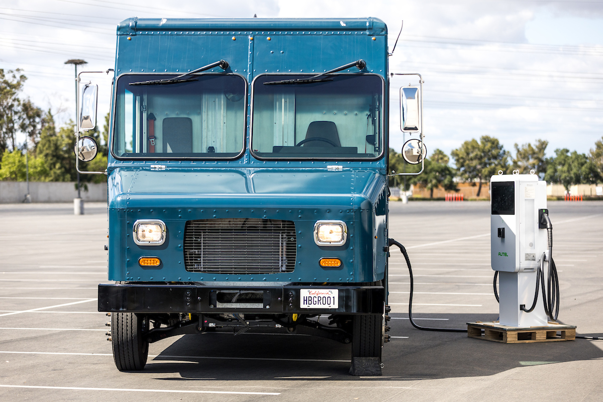 Blue EV step van facing forward, with charger to the right.